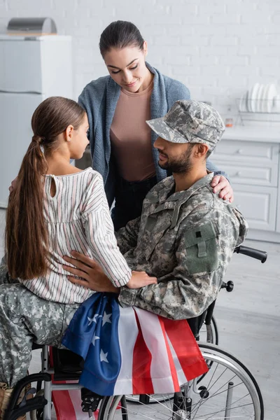 Mulher e criança olhando para o pai em uniforme militar e cadeira de rodas com bandeira americana em casa — Fotografia de Stock