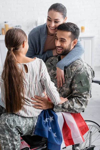 Hombre sonriente en uniforme militar y silla de ruedas con bandera americana mirando al niño cerca de su esposa en casa - foto de stock