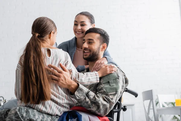 Homem de uniforme militar e cadeira de rodas com bandeira americana sorrindo para a filha perto da esposa em casa — Fotografia de Stock