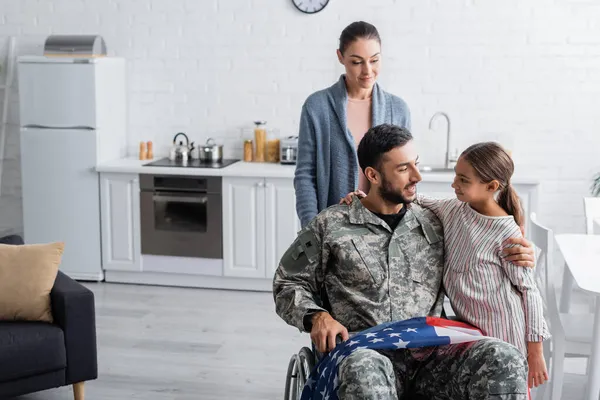 Veterano sonriente en silla de ruedas con bandera americana abrazando a su hija cerca de su esposa en casa - foto de stock