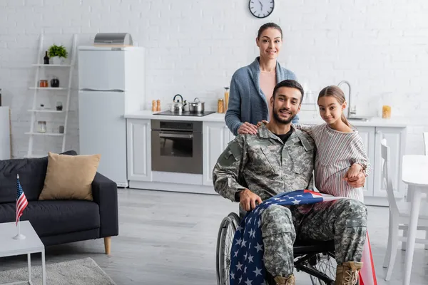 Famille regardant la caméra près du père en uniforme militaire et fauteuil roulant avec drapeau américain à la maison — Photo de stock