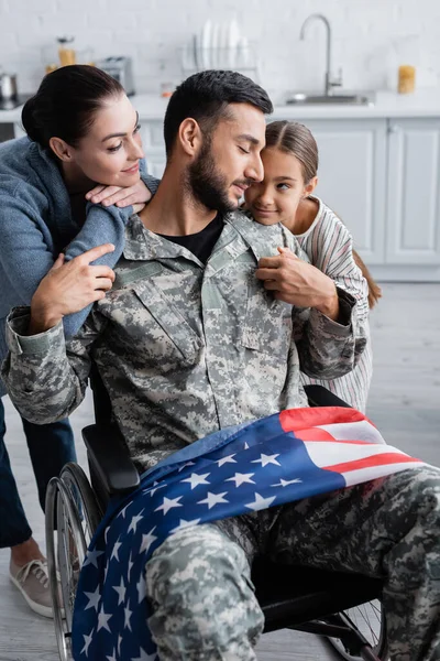 Soldado em cadeira de rodas com bandeira americana perto de filha e esposa em casa — Fotografia de Stock