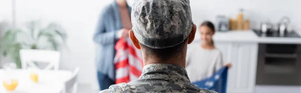 Hombre en uniforme militar cerca de la familia borrosa con bandera americana en casa, pancarta - foto de stock