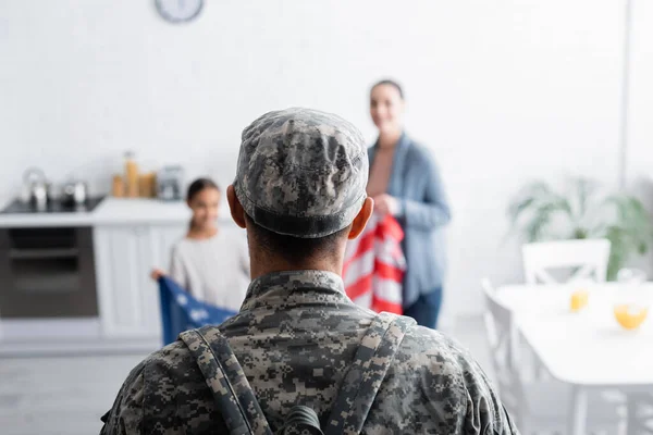 Soldat en camouflage debout près de la famille floue avec le drapeau américain à la maison — Photo de stock