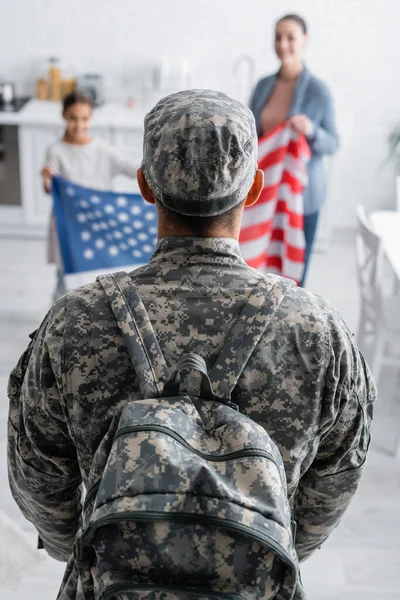 Mann in Tarnuniform mit Rucksack neben verschwommener Familie mit amerikanischer Flagge zu Hause — Stockfoto