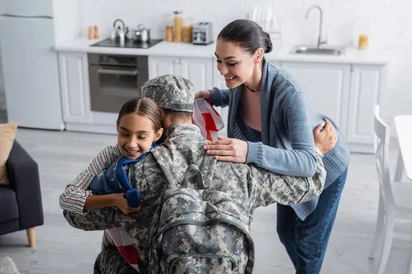 Donna sorridente che tiene bandiera americana mentre figlia abbraccia il padre in uniforme militare a casa — Foto stock