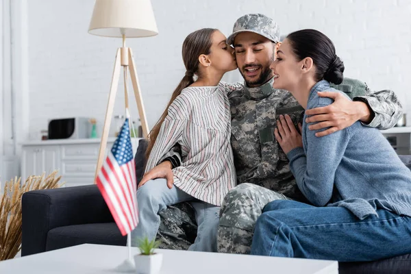 Kid beijando pai em uniforme militar perto de mãe e bandeira americana em casa — Fotografia de Stock