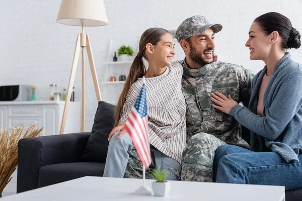 Felice famiglia abbracciare l'uomo in uniforme mimetica sul divano vicino bandiera americana a casa — Foto stock