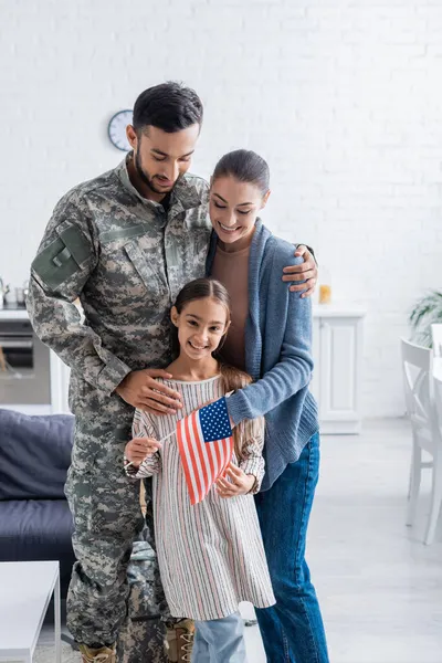 Sorrindo homem em uniforme militar abraçando esposa e filho com bandeira americana em casa — Fotografia de Stock