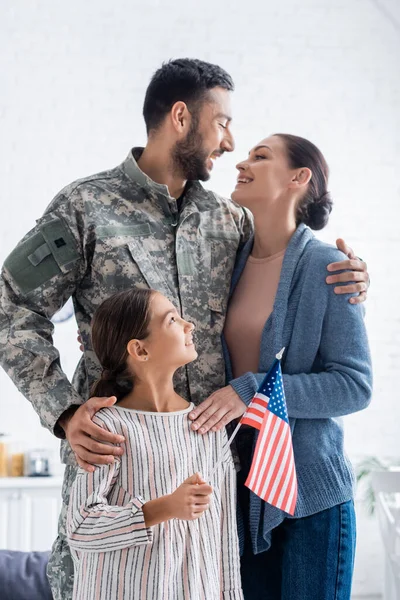 Mulher positiva olhando para o marido em uniforme de camuflagem perto da filha com bandeira americana em casa — Fotografia de Stock