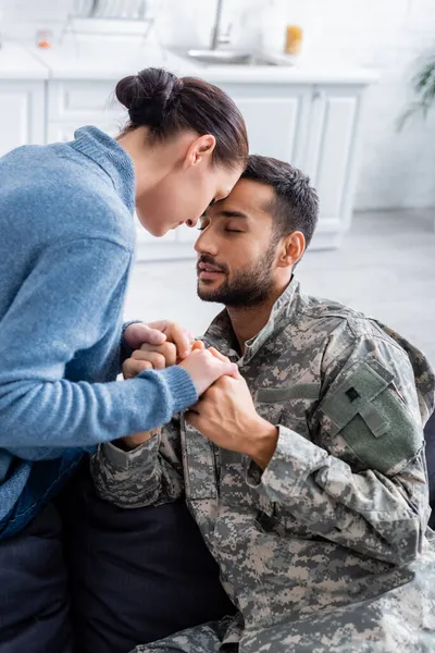 Mujer cogida de la mano del marido en uniforme de camuflaje en casa - foto de stock