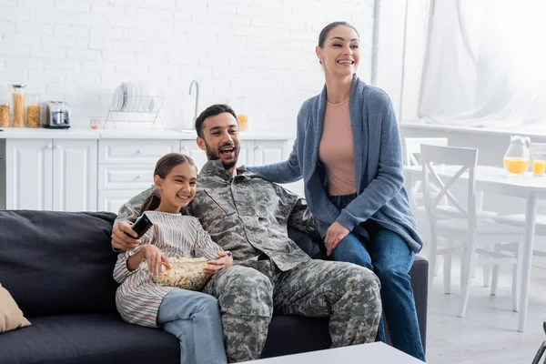 Sonriente hombre en uniforme militar sosteniendo mando a distancia cerca de la familia con palomitas de maíz en casa - foto de stock