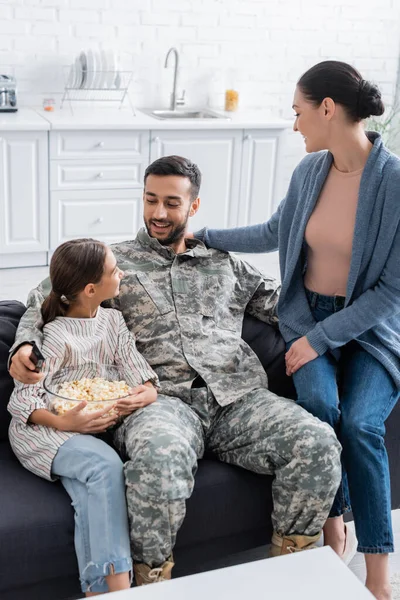 Soldado sosteniendo mando a distancia cerca de hija con palomitas de maíz y esposa en casa - foto de stock