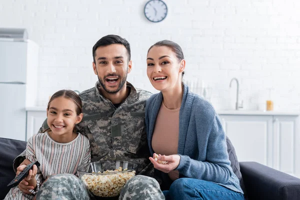Hombre positivo en uniforme militar sentado cerca de la familia con mando a distancia y palomitas de maíz en el sofá - foto de stock
