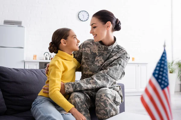 Vista lateral da mãe em uniforme militar abraçando criança perto da bandeira americana na mesa em casa — Fotografia de Stock