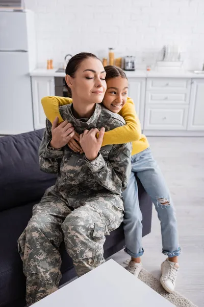 Sonriente niño preadolescente abrazando a la madre en uniforme militar en el sofá - foto de stock