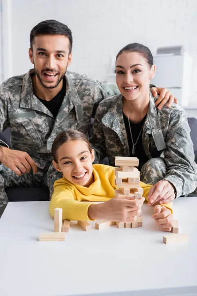 Genitori felici in uniforme militare guardando la fotocamera vicino al bambino con blocchi di legno gioco a casa — Foto stock