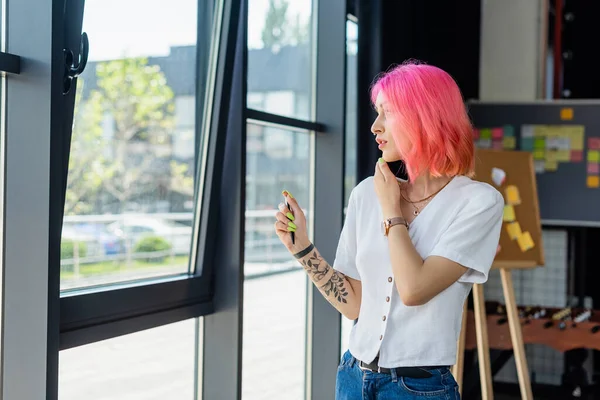 Молодая деловая женщина с розовыми волосами разговаривает на смартфоне, глядя в окно — стоковое фото
