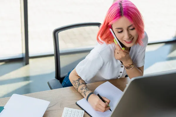 Femme d'affaires joyeuse avec les cheveux roses parler sur smartphone et écrire sur ordinateur portable près de moniteur d'ordinateur — Photo de stock