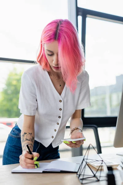 Проколота молода бізнес-леді з рожевим волоссям тримає смартфон і пише в блокноті — стокове фото