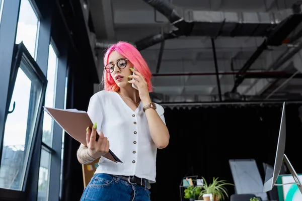 Low angle view of pierced manager with pink hair talking on cellphone and looking at folder — Stock Photo