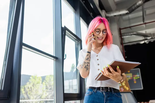 Gérant heureux et percé avec les cheveux roses parlant sur téléphone portable et regardant dossier — Photo de stock