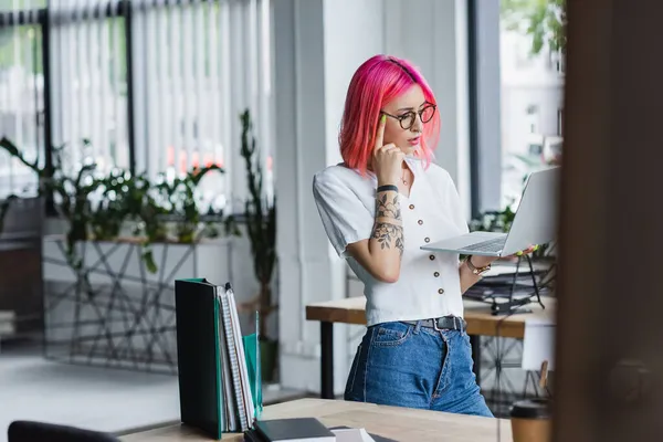 Задумчивая молодая деловая женщина с розовыми волосами, держащая ноутбук в офисе — стоковое фото