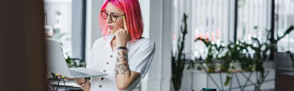 Стомлена молода бізнес-леді з рожевим волоссям тримає ноутбук в офісі, банер — стокове фото
