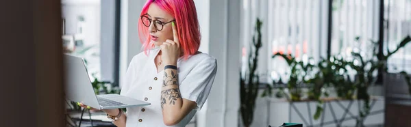 Pensativo joven mujer de negocios con el pelo rosa que sostiene el ordenador portátil en la oficina, bandera - foto de stock