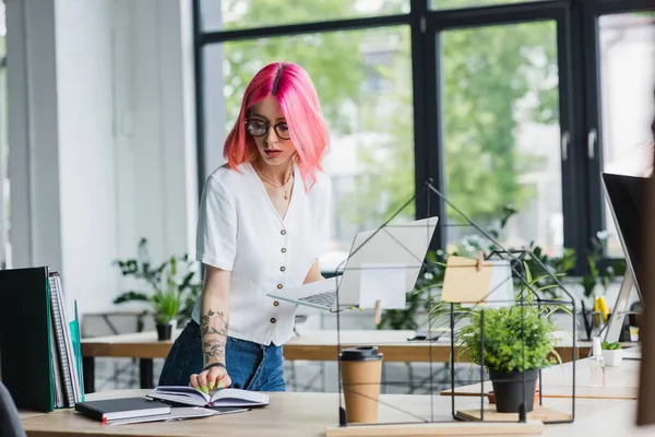 Junge Geschäftsfrau mit rosa Haaren hält Laptop in der Hand und schaut auf Notizbuch — Stockfoto