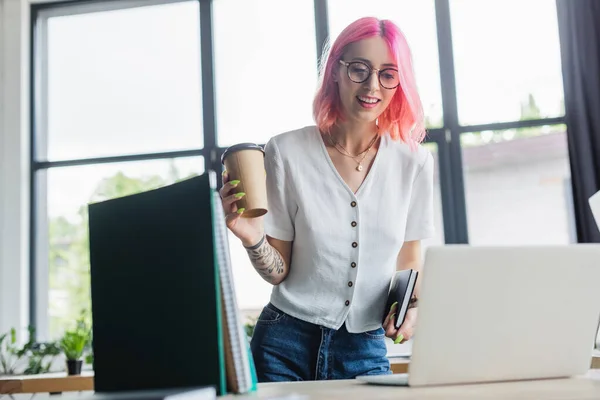 Fröhlicher und gepiercter Manager mit rosa Haaren, Pappbecher in der Hand und Blick auf Laptop — Stockfoto