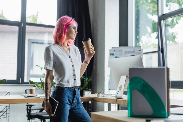 Vista lateral de mujer de negocios perforada con pelo rosa sosteniendo taza de papel y cuaderno - foto de stock