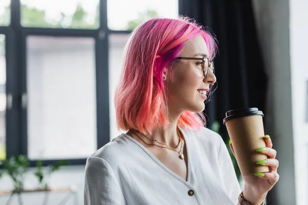 Visão lateral de empresária perfurada com cabelo rosa segurando copo de papel e sorrindo no escritório — Fotografia de Stock
