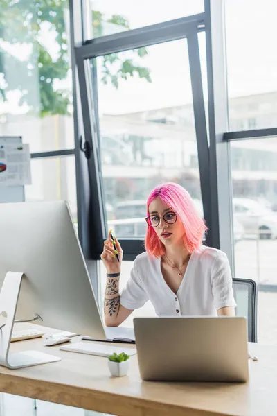 Gepiercte Geschäftsfrau mit pinkfarbenen Haaren hält Stift in der Hand und blickt auf Computermonitor — Stockfoto