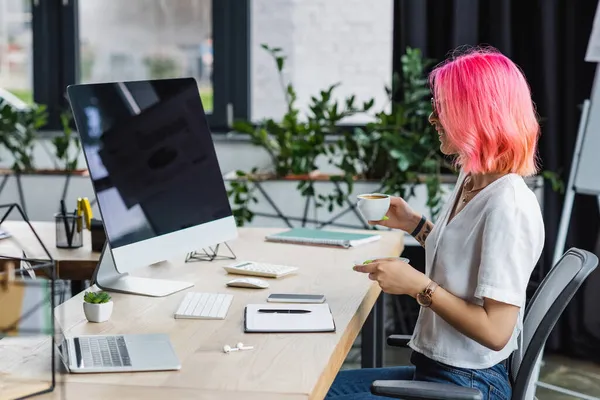 Vista lateral de mulher de negócios feliz com cabelo rosa segurando xícara de café e olhando para monitor de computador — Fotografia de Stock