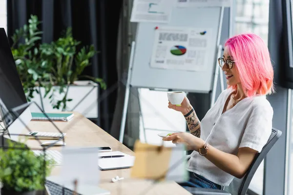 Femme d'affaires joyeuse avec les cheveux roses tenant tasse de café et regardant moniteur d'ordinateur — Photo de stock