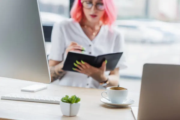 Kaffeetasse in Pflanzennähe und verschwommene Geschäftsfrau mit Notizbuch — Stockfoto