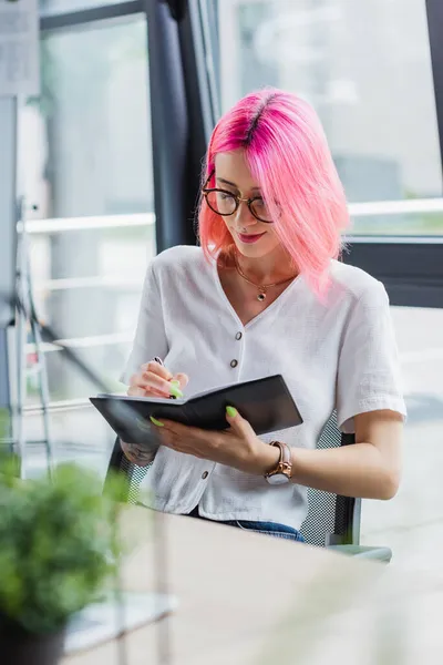Tätowierte Geschäftsfrau mit rosa Haaren schreibt auf Notizbuch — Stockfoto