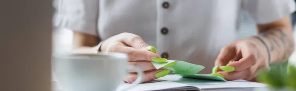 Cropped view of tattooed businesswoman holding sticky note near notebook and blurred cup, banner — Stock Photo