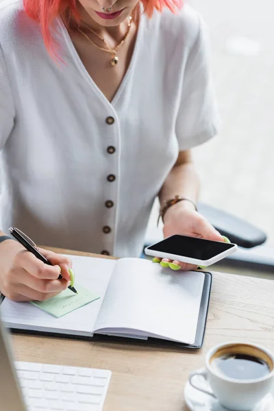 Vue recadrée d'une femme d'affaires percée écrivant sur une note collante et tenant un smartphone avec écran vierge — Photo de stock