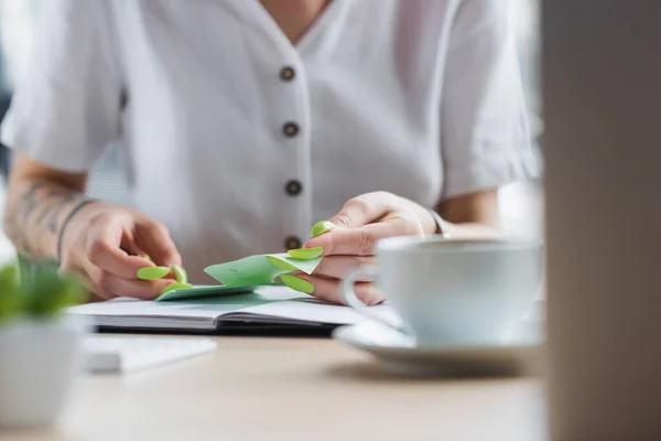 Cropped view of tattooed businesswoman holding sticky note near notebook and blurred cup — Stock Photo