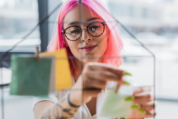 Femme d'affaires souriante aux cheveux roses tenant une épingle en bois près d'une note floue sur un panneau métallique — Photo de stock