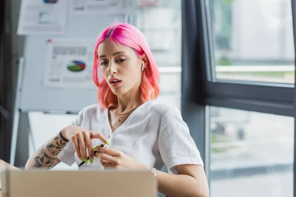 Pierced businesswoman in earphone holding pen and looking at laptop — Stock Photo