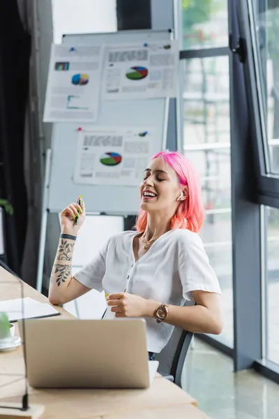 Glückliche Geschäftsfrau mit Kopfhörer hält Stift in der Hand und lacht neben Laptop — Stockfoto