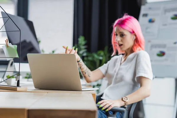 Ordinateur portable près floue femme d'affaires avec les cheveux roses gestuelle au bureau — Photo de stock