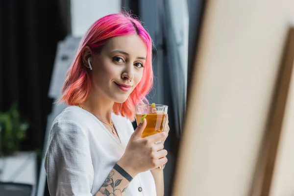 Femme d'affaires joyeuse avec les cheveux roses et écouteur tenant tasse de thé au bureau — Photo de stock