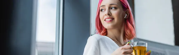 Lächelnde Geschäftsfrau mit rosafarbenen Haaren und Kopfhörer, die eine Tasse Tee im Büro hält, Banner — Stockfoto