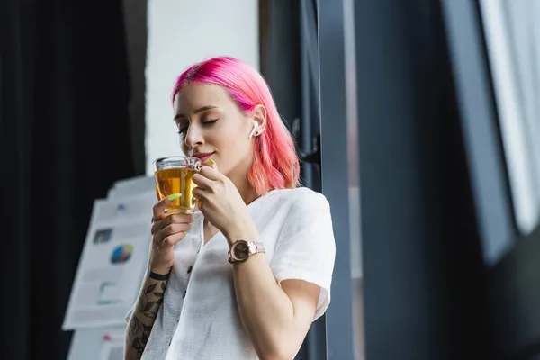 Довольная деловая женщина с розовыми волосами и наушником, нюхающая чашку чая в офисе — стоковое фото
