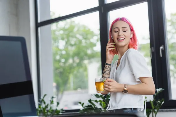 Femme d'affaires heureuse avec cheveux roses et écouteurs sans fil tenant tasse de thé au bureau — Photo de stock