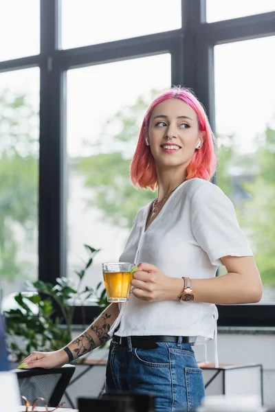 Joyeuse femme d'affaires avec des cheveux roses et écouteurs tenant tasse de thé au bureau — Photo de stock